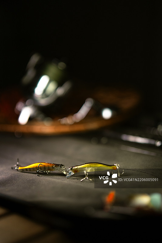 鲜艳的钓鱼饵和渔具箱的特写，背景中有钓鱼卷线器和渔网，色调较暗图片素材