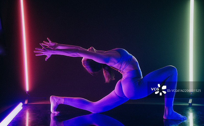 在黑暗的房间里，女人在反射地板上做瑜伽姿势，周围有五彩斑斓的霓虹灯。图片素材