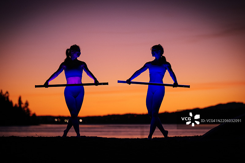 两位女性在华盛顿州贝恩布里奇岛的湖边日落时分练习武术和舞蹈，手持长棍。图片素材