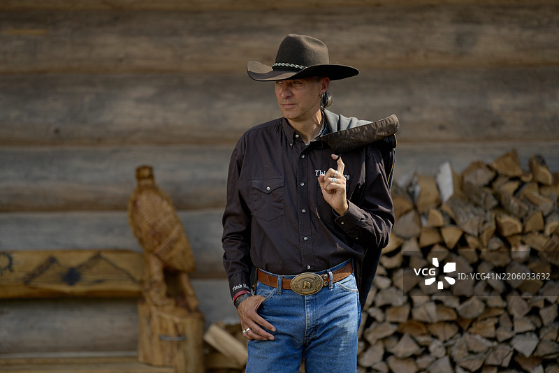 穿着黑色衬衫和帽子的牛仔站在一座木屋旁，旁边有一堆柴火。德国布兰登堡图片素材