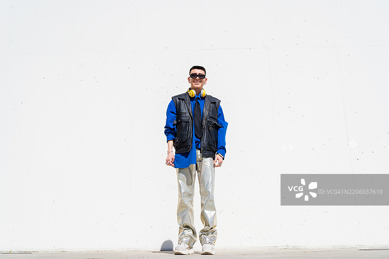 时尚年轻男子全身肖像，靠在白色墙壁上，穿着银色裤子、蓝色衬衫和黑色背心。图片素材