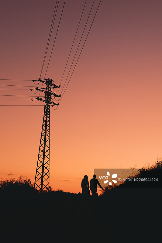 在乡村地区和电塔附近，夕阳下行走的一对情侣剪影。图片素材