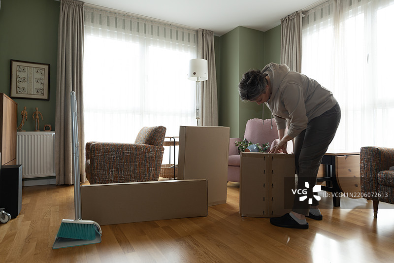 独自一人在空荡荡的公寓房间里搬家的活跃老年女性正在从纸箱中拆包。图片素材