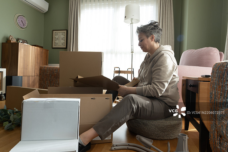 独自一人在空荡荡的公寓房间里搬家的活跃老年女性正在从纸箱中拆包。图片素材