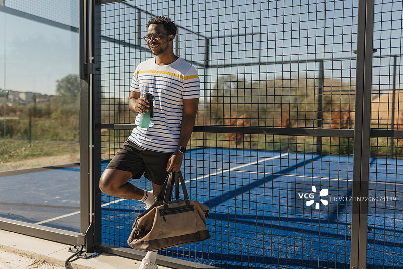 男子在球场入口处放松，身旁有网球装备图片素材