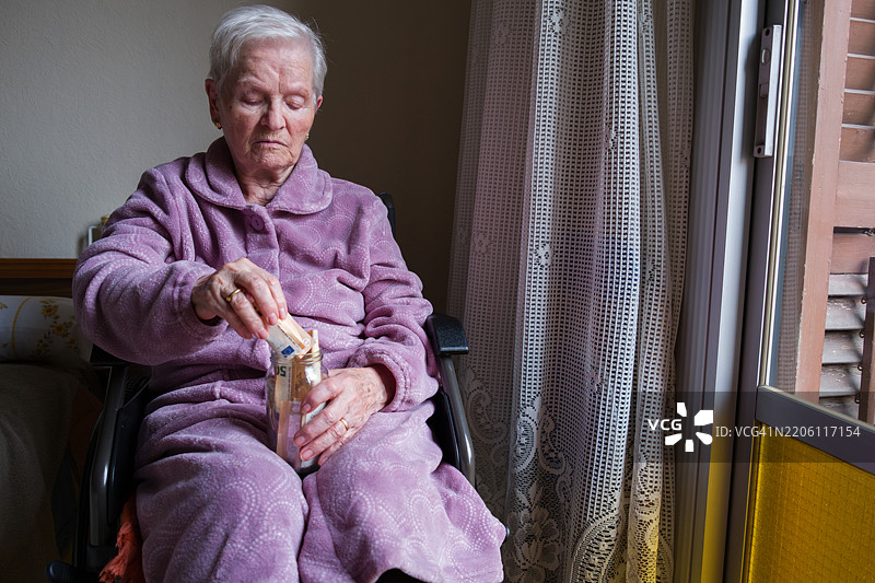 一位年长女性在家中将欧元纸币放入罐子里图片素材