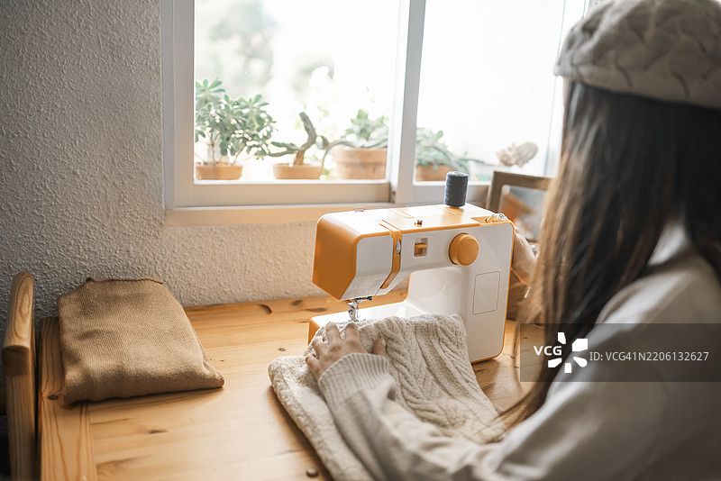 女裁缝在桌子上用现代缝纫机缝制衣服，阳光透过窗户洒进来，窗边有植物。图片素材