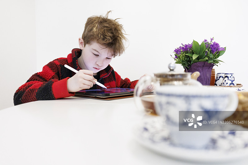 男孩使用数字平板电脑和触控笔。亚洲孩子在白色桌子上绘画，桌上有茶杯和花朵。图片素材