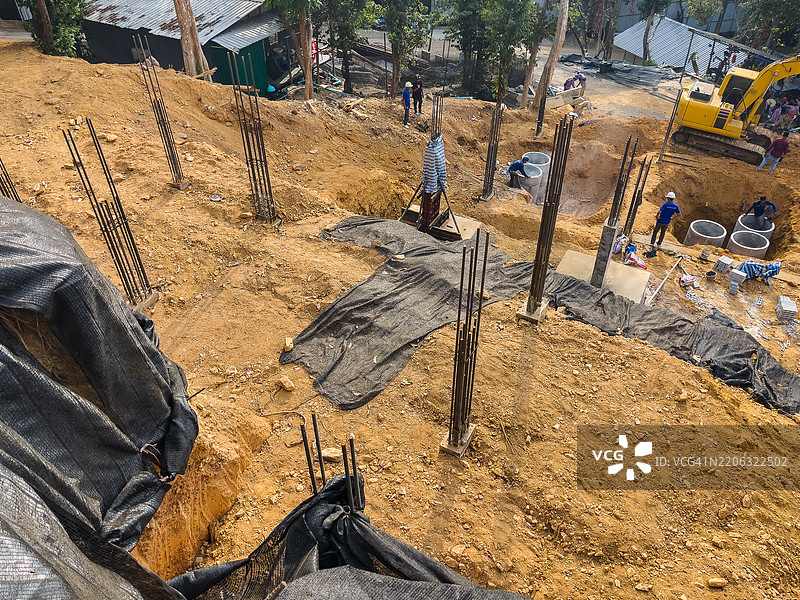 建筑工地上有挖掘机、工人、钢筋和正在进行的基础工程，周围是树木环绕的丘陵地形。图片素材