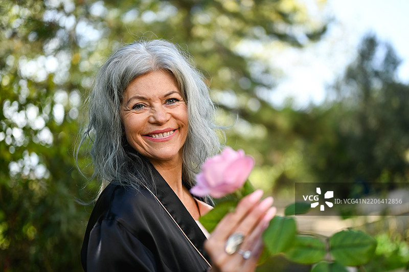 美丽微笑的成熟的女性在花园中展示一朵粉色玫瑰图片素材