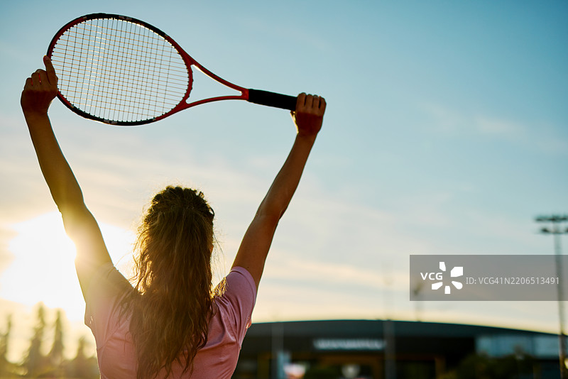 女性网球运动员在日落时分举起双臂和网球拍庆祝获胜图片素材