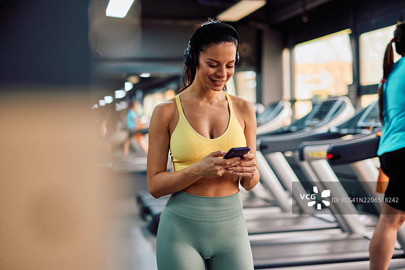 微笑的运动员在健身房使用手机并佩戴耳机图片素材