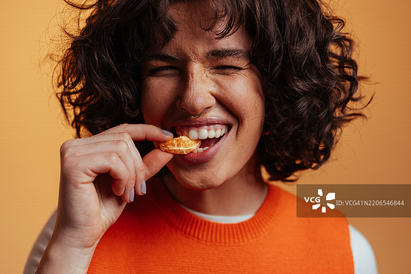 年轻女子闭着眼睛吃着一片橘子，卷曲的头发随风飘扬。图片素材