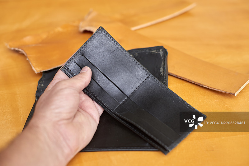 熟练的皮革工匠在工作台旁拿着手工制作的黑色钱包，展示传统工艺，周围散落着皮革碎片，放在风化的木质表面上。图片素材