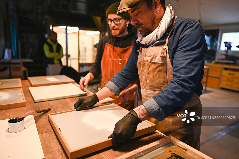 两名穿着围裙和手套的男性印刷工人在他们的工作室里准备丝网印刷框架图片素材