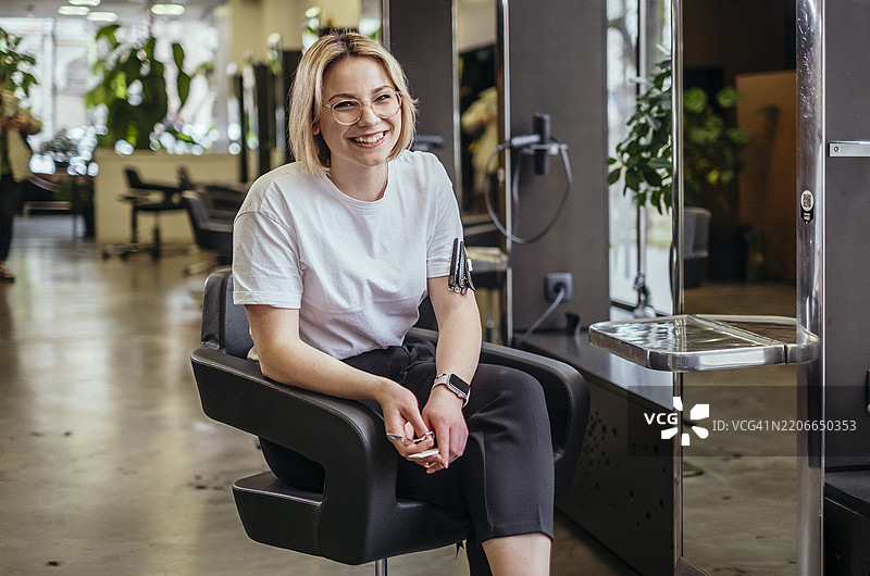 年轻女性美容院老板在现代化的发廊中微笑，阳光透过窗户洒入，配备专业设备。图片素材