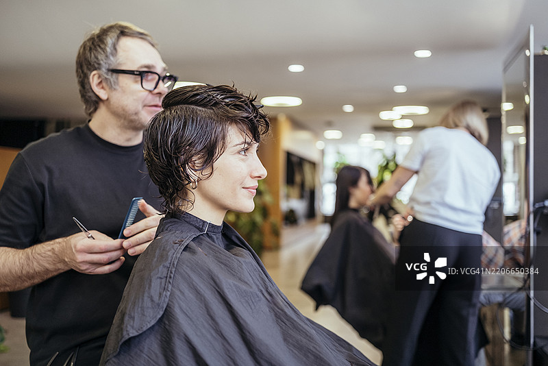 中年男性发型师在现代沙龙为微笑的女性客户修剪短发图片素材