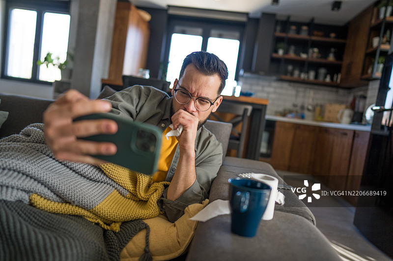 生病的男人在沙发上咳嗽时使用手机图片素材