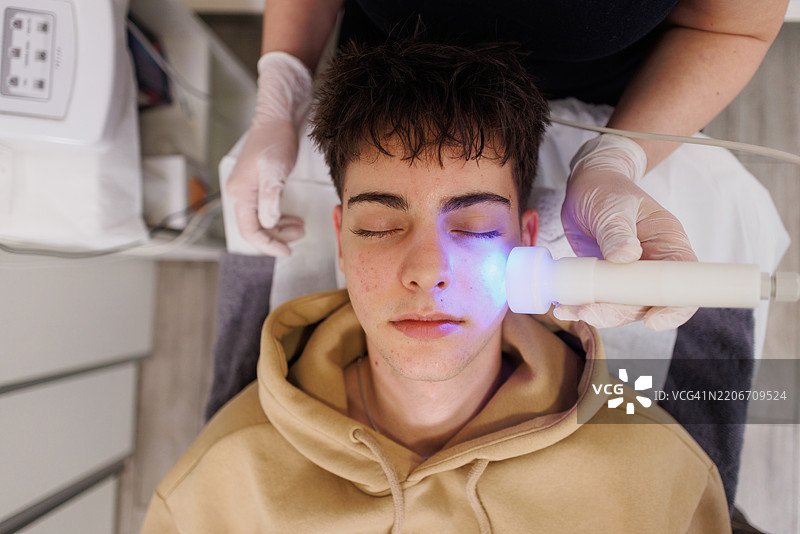 美容师为青少年男孩的脸部进行LED光疗图片素材