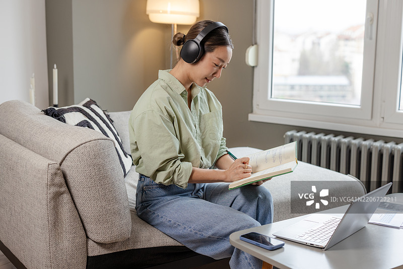 亚洲女性戴着耳机在家用笔记本电脑写笔记图片素材