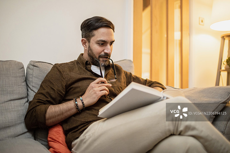 一个男人坐在沙发上，嘴里叼着眼镜看书。图片素材