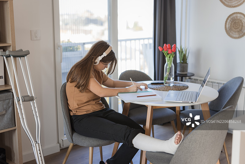 因受伤而在家进行在线学习的女学生，佩戴耳机并使用笔记本电脑。图片素材