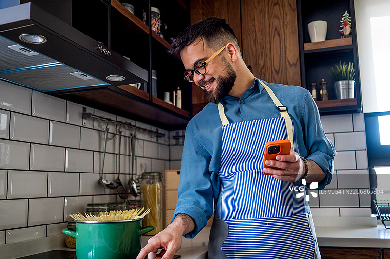 一位年轻男子在厨房里，穿着蓝色围裙，仔细地按照手机上的食谱煮意大利面。图片素材