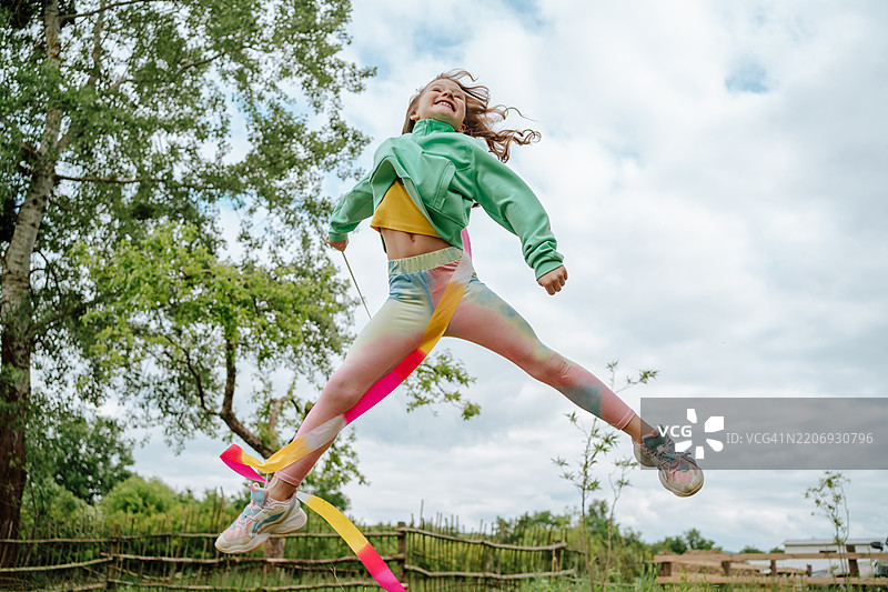 空中跳跃的彩带体操运动员。微笑的小女孩在大自然中跳跃，手中挥舞着五彩缤纷的彩带。图片素材