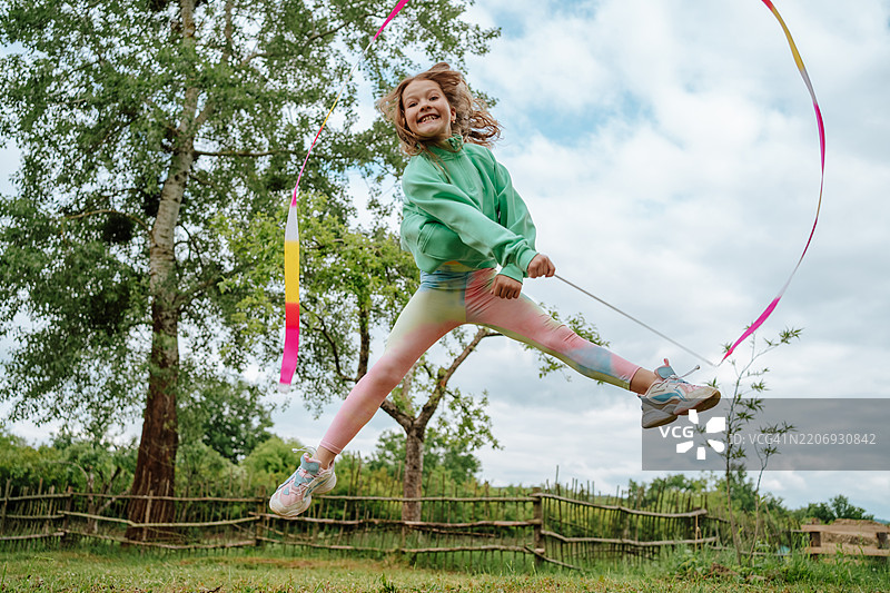 快乐的调皮女孩在大自然中跳起舞来，手中挥舞着五彩缤纷的艺术体操彩带。图片素材