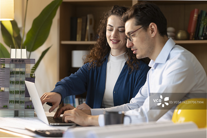 两位建筑师在办公室合作进行可持续建筑项目，使用笔记本电脑和比例模型。图片素材