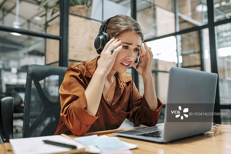 呼叫中心的客服专员坐在桌前，按压太阳穴，担忧地看着笔记本电脑。图片素材