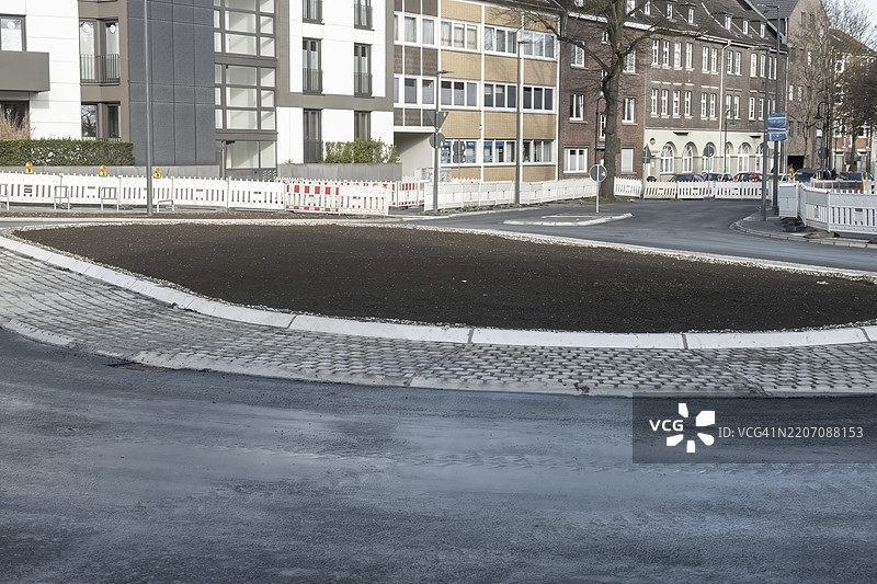 新铺好的土壤位于城市环境中新建的交通岛上，周围是建筑物和施工围栏，等待绿化，德国北莱茵-威斯特法伦州博特罗普。图片素材