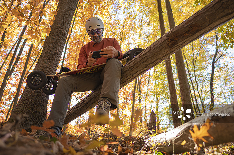 秋季森林中，坐在倒下的树木上，使用手机的电动山地滑板男士图片素材