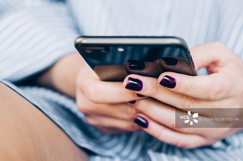 女性使用手机输入文本，查看社交媒体或在线购物。特写镜头展示了涂有紫色指甲油的女性手握着一部金色智能手机。她坐在一张木桌前，穿着条纹蓝色衬衫。图片素材