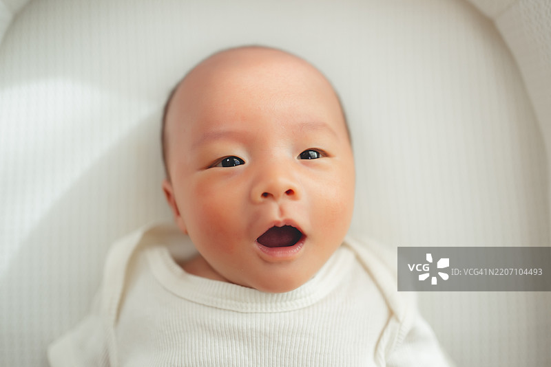 亚洲新生儿穿着连体衣躺在客厅的白色床上。图片素材