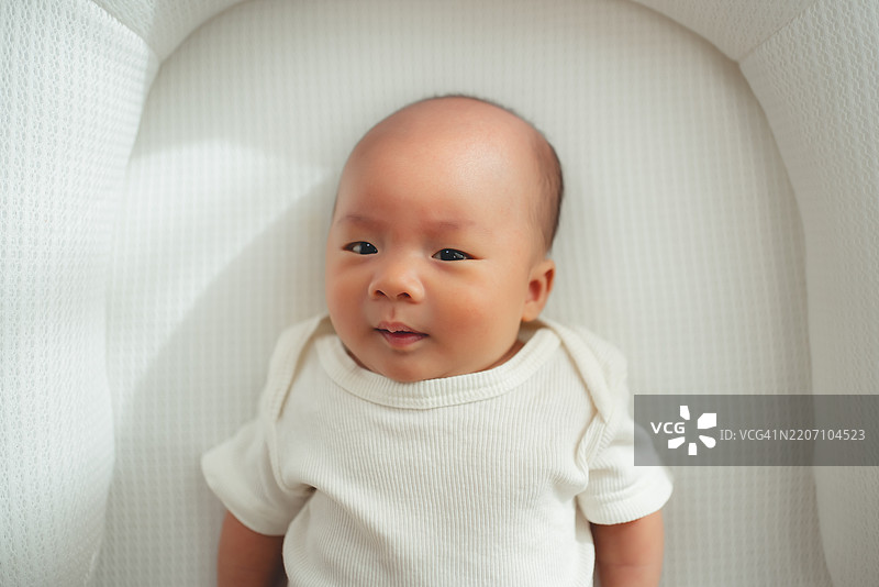 亚洲新生儿穿着连体衣，躺在客厅的白色床上。图片素材