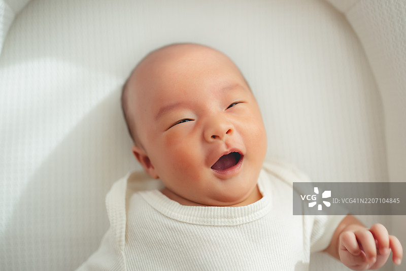 亚洲新生儿穿着连体衣躺在客厅的白色床上。图片素材