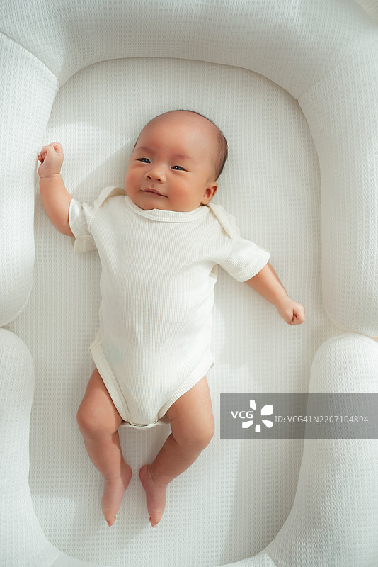 亚洲新生儿穿着连体衣躺在客厅的白色床上。图片素材