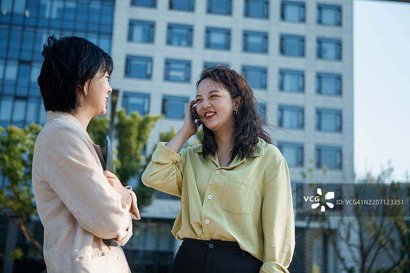 两位女性在办公大楼前手持文件讨论工作 - 合作、商业、贸易、投资、工作、职业图片素材