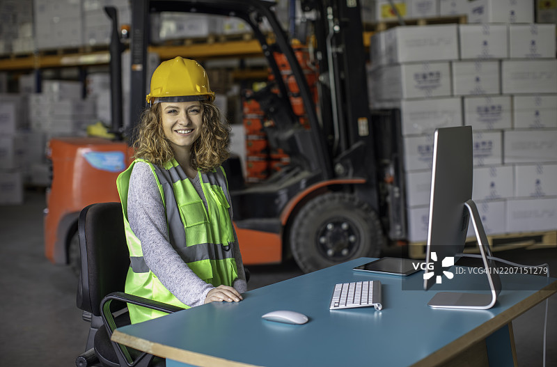 微笑的女性仓库员工在工业工作场所的桌子旁，戴着黄色安全帽和安全背心。图片素材