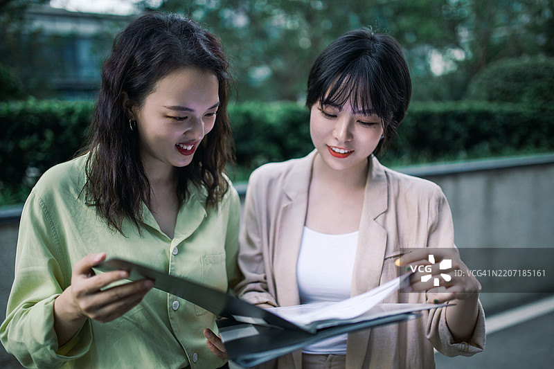 两位女性商务人士在商业办公大楼公园中交流，手中拿着咖啡和文件。图片素材