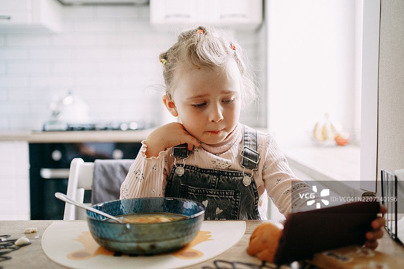 小女孩在厨房吃午餐时佩戴着人工耳蜗，专注地看着智能手机图片素材