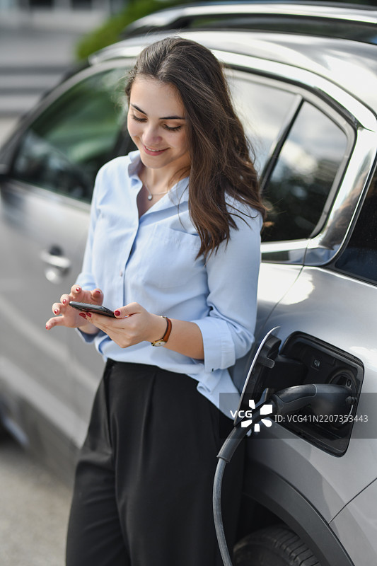 快乐的女性在给电动车充电时使用智能手机，靠在车上。图片素材