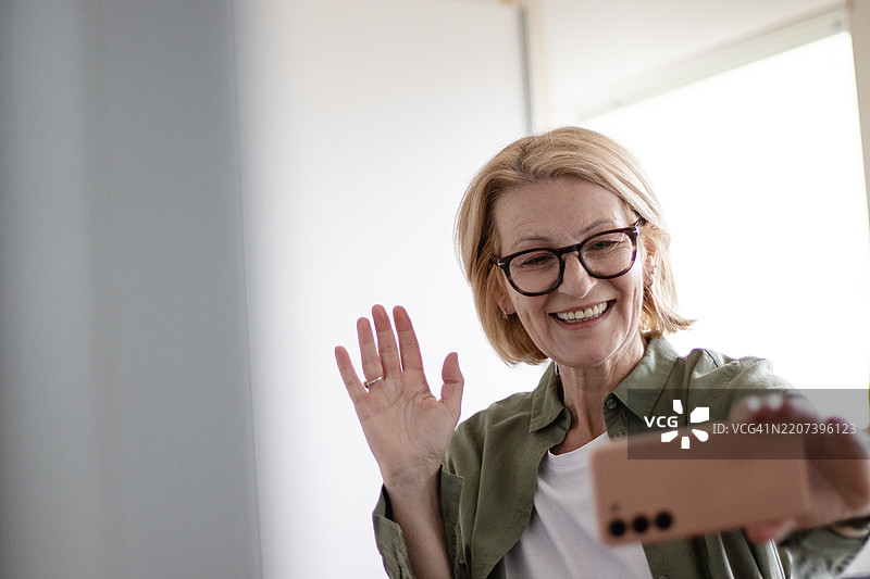 成熟的金发快乐女戴着眼镜，坐在桌子前用手机进行视频通话。图片素材