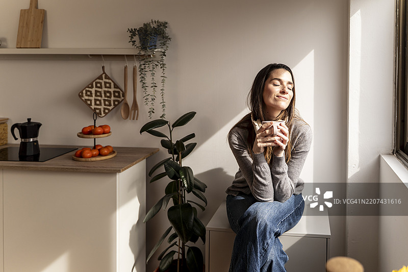 宁静的女人在温暖的早晨光线中，悠闲地品尝热饮，坐在舒适的厨房环境中。图片素材
