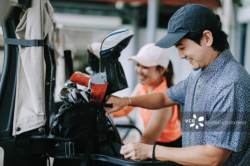亚洲华裔男性高尔夫球手和朋友在高尔夫球场的周末早晨将高尔夫球包放上高尔夫球车图片素材