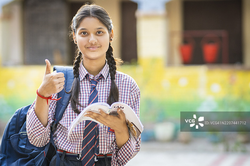 快乐的女学生穿着校服，手里拿着一本书，站在外面微笑并竖起大拇指。图片素材