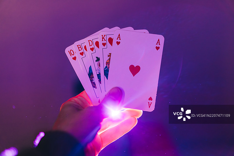 手中持有从10到A的扑克牌，背景昏暗，霓虹灯光。皇家同花顺组合。图片素材
