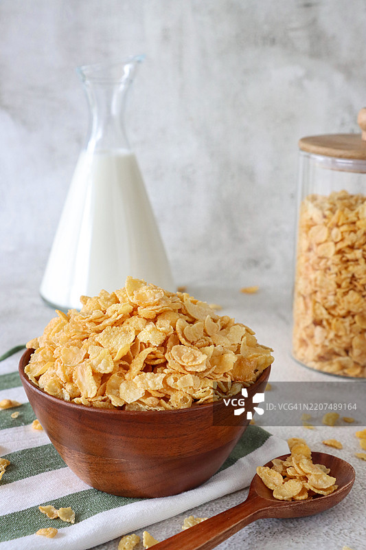 特写镜头，木碗里装满了脆脆的烤玉米片，放在绿色和白色条纹的茶巾上，旁边有木制汤勺、玻璃储存罐和牛奶壶，背景为斑驳的灰白色，焦点在前景。图片素材
