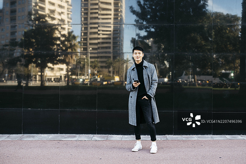 西班牙，巴塞罗那，微笑的年轻男子，穿着黑色高领毛衣和灰色外套，手持手机图片素材
